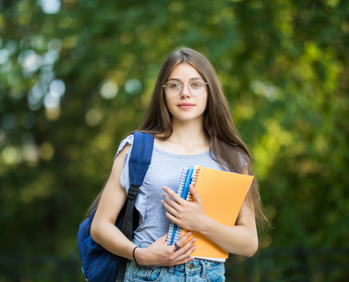 Estudante de óculos e cabelos compridos com mochila no ombro, segurando cadernos