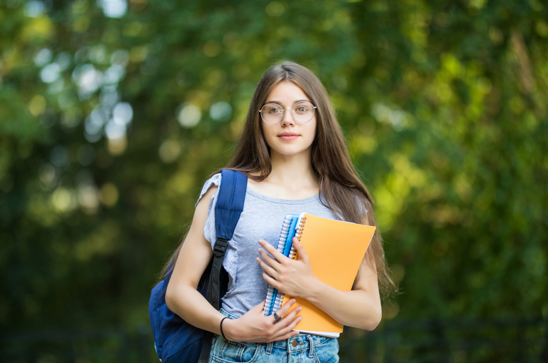 Estudante de óculos e cabelos compridos com mochila no ombro, segurando cadernos