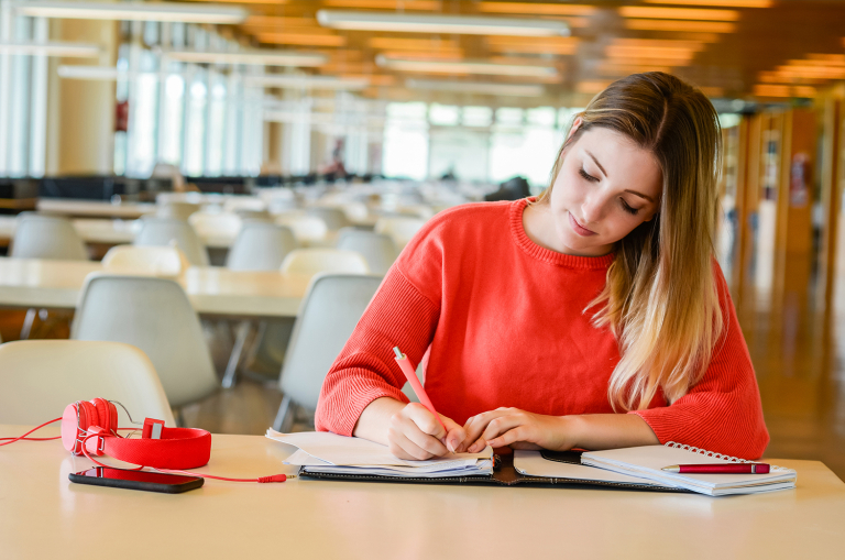 Estudante de blusa vermelha escrevendo em caderno na biblioteca